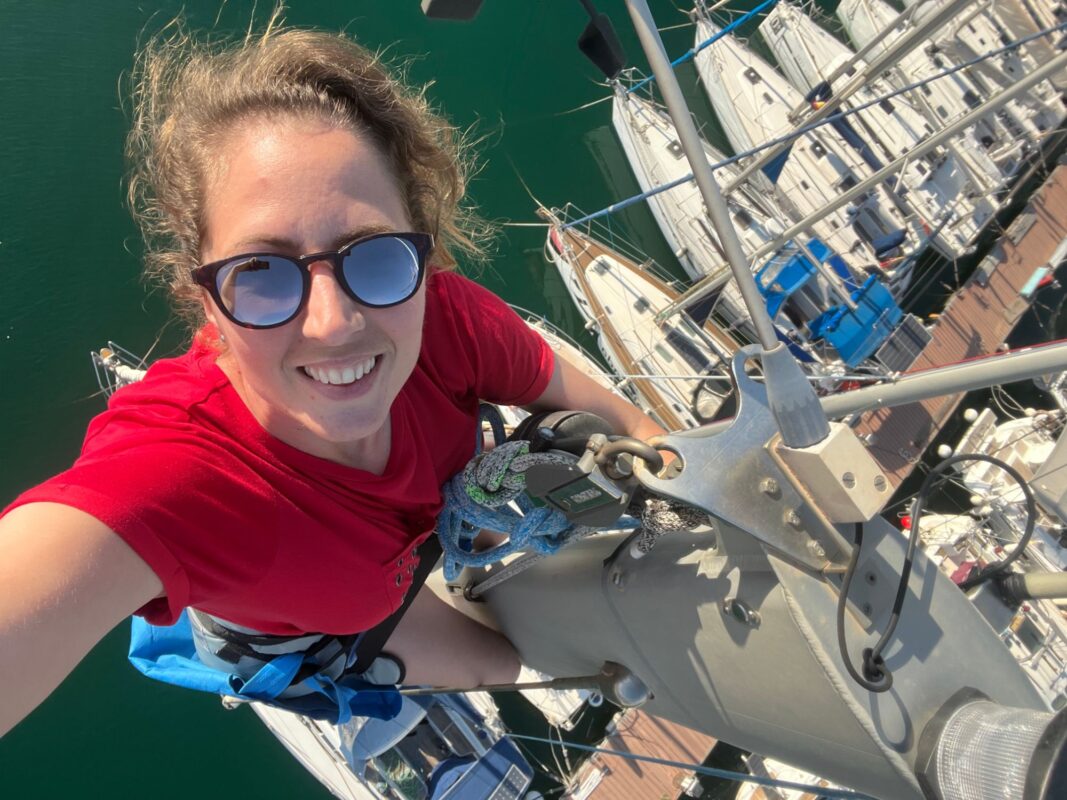 Elena Oceánide sonriendo en lo alto del mástil de su velero, con los barcos del puerto debajo